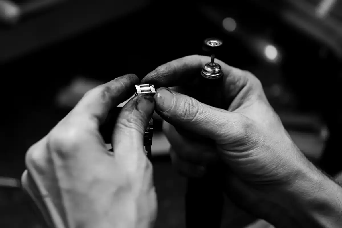 Macro of two working hands steadying and polishing a small bright metal component over a dark bench.