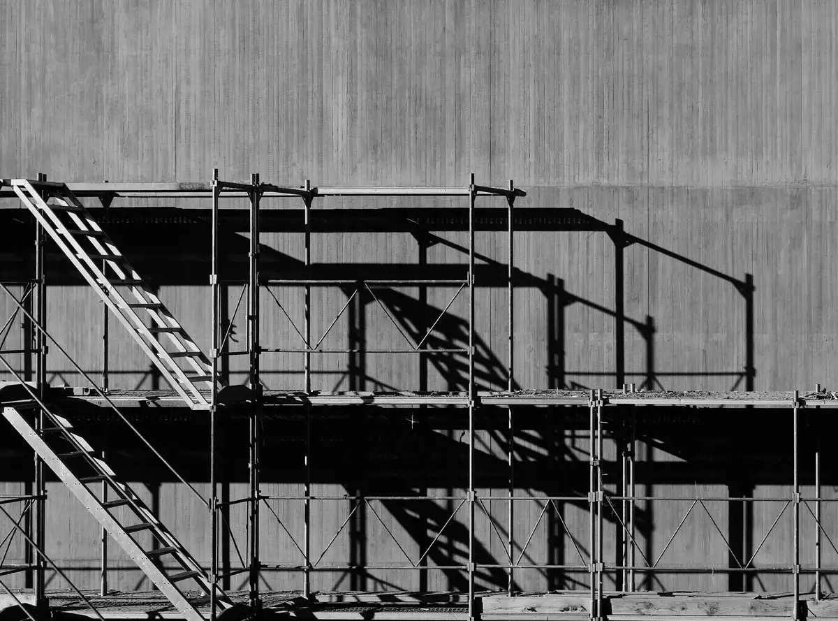 Scaffolding tiers and a stair run standing off a board-marked concrete wall in hard midday sun.