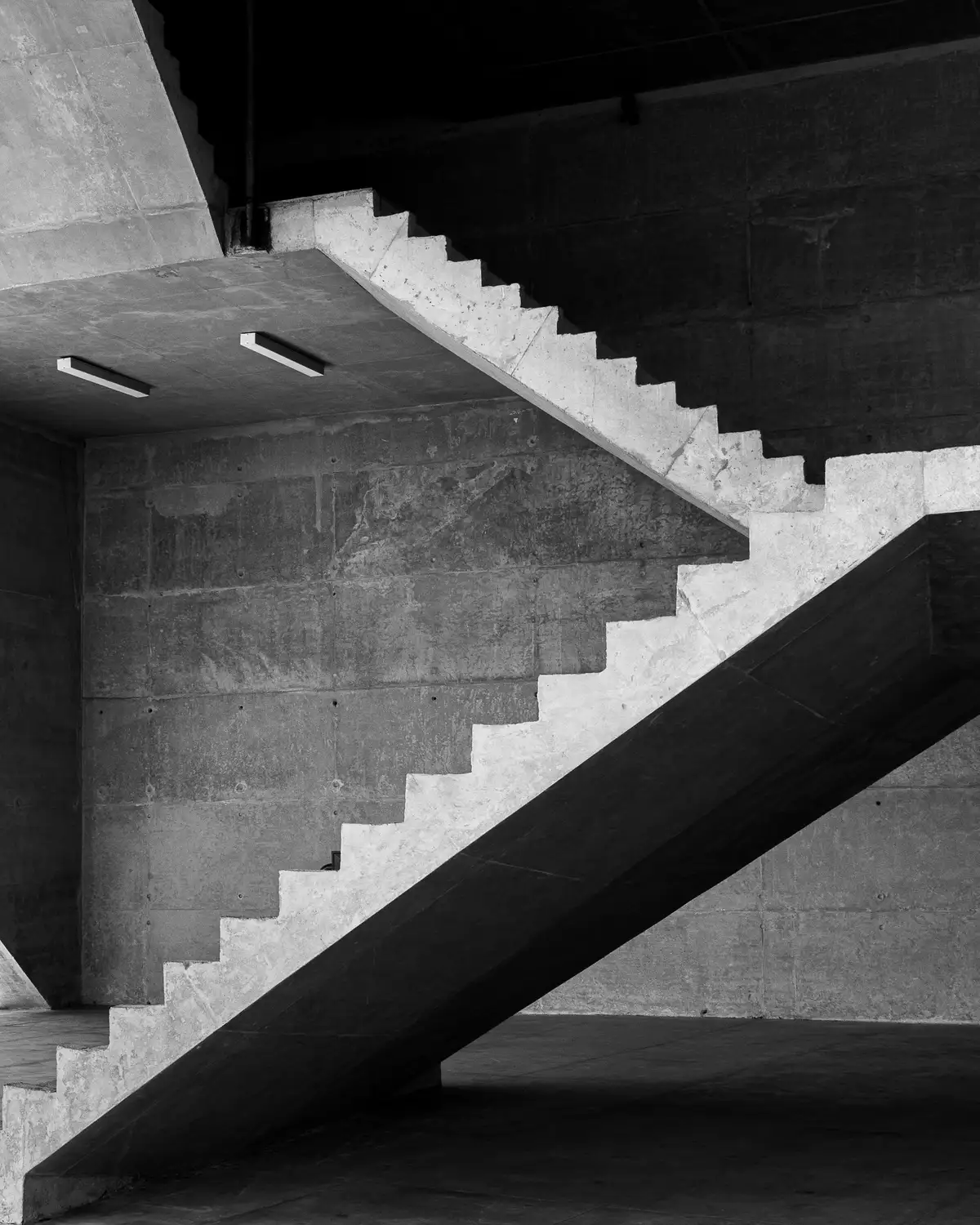 A raw concrete stair flight cutting a bright zigzag diagonal across a board-marked wall in deep shadow.