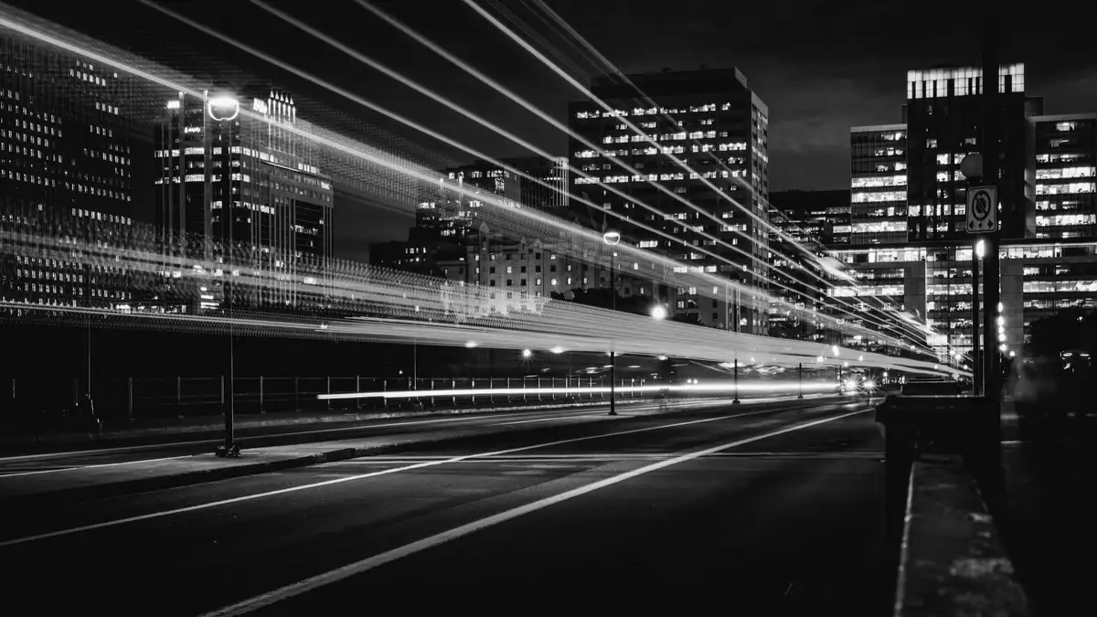 Night long-exposure of an empty roadway: continuous horizontal light trails streak past dark buildings.