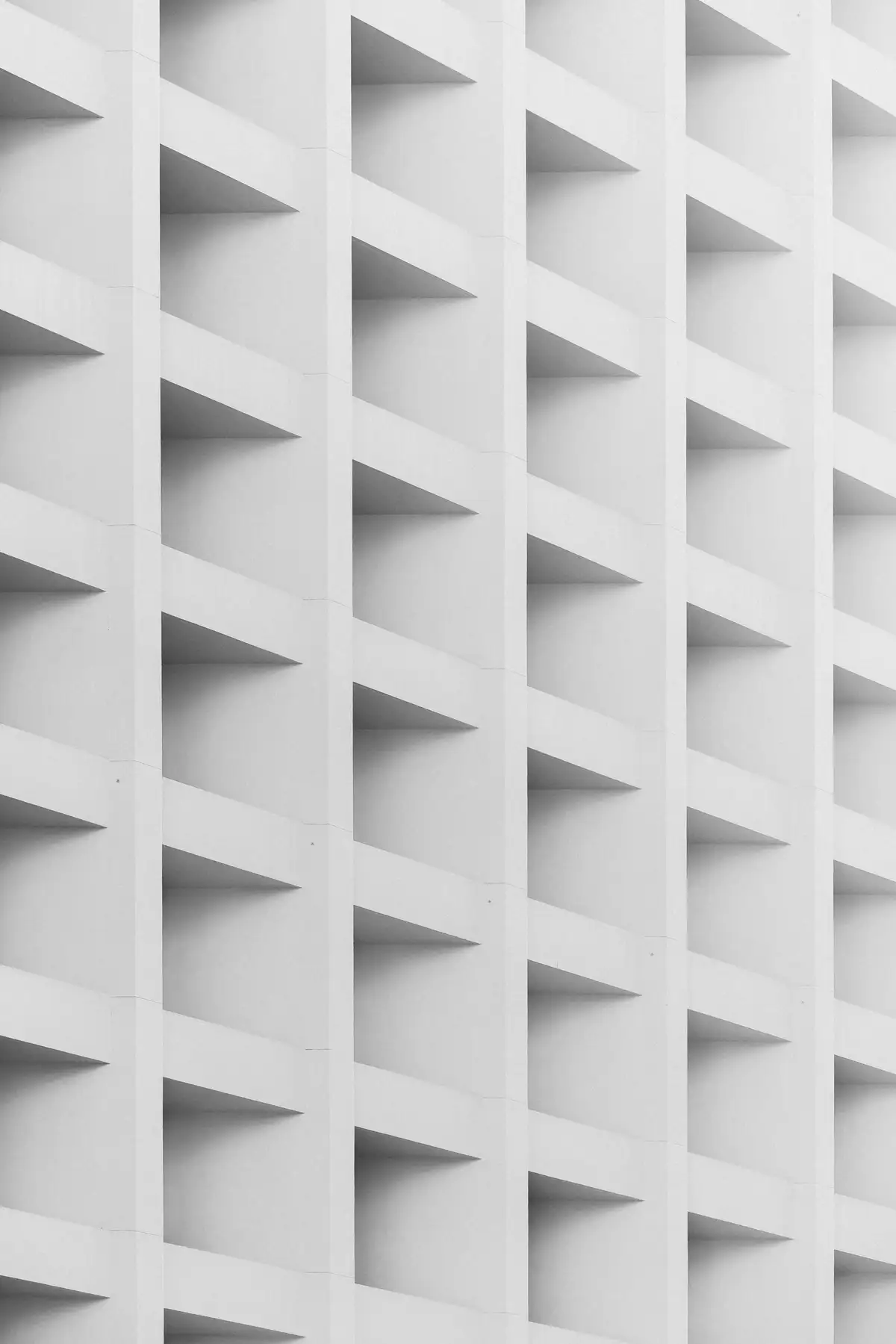 Flat-on view of a white concrete facade: an orthogonal grid of deep square recesses, each casting a hard triangular shadow.